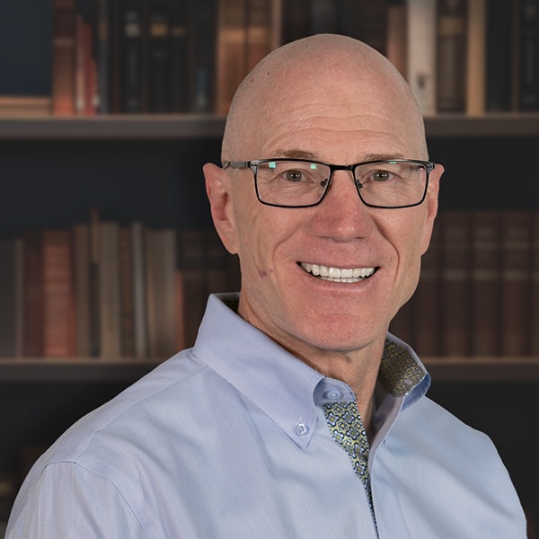 A professional headshot of a smiling, bald man with glasses, wearing a light blue button-down shirt in front of a bookshelf.