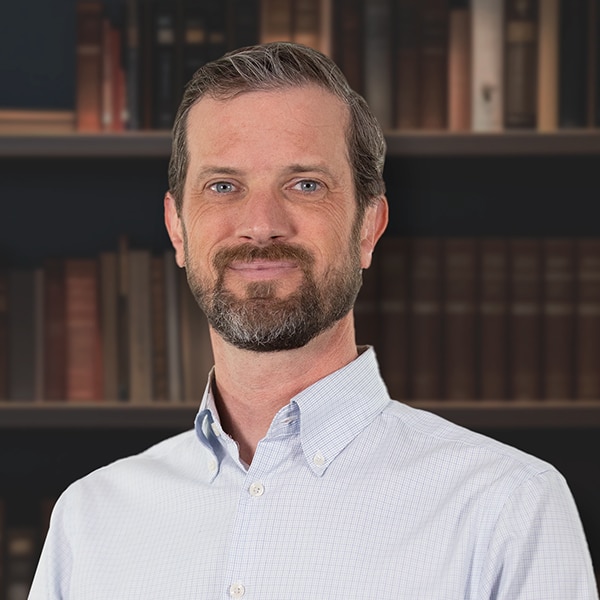 A professional headshot of a smiling man with a beard and blue eyes, wearing a light blue patterned button-down shirt in front of a bookshelf.