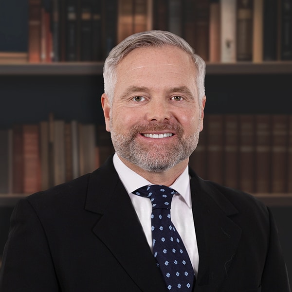 A professional headshot of a smiling man with a gray beard, wearing a black suit and a patterned tie, in front of a bookshelf.