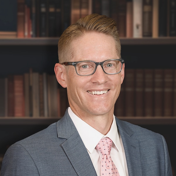 A professional headshot of a smiling man with glasses, wearing a grey suit and a pink patterned tie, in front of a bookshelf.