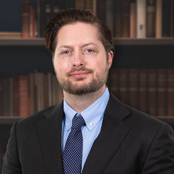 A professional headshot of a man with a beard, wearing a dark suit, blue shirt, and patterned tie in front of a bookshelf.